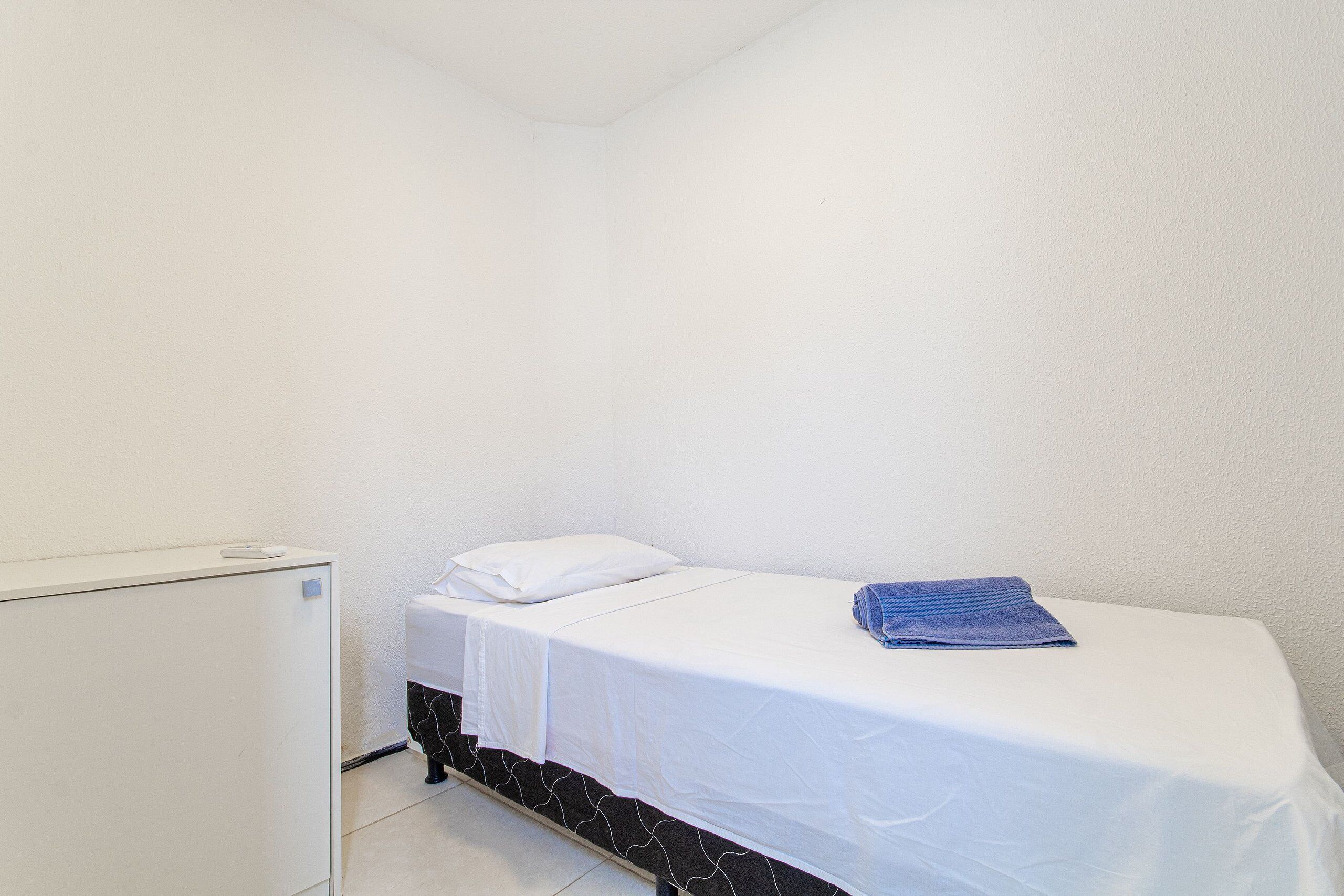 A small, single-bed room with a white bed, a white wall, and a white cabinet.