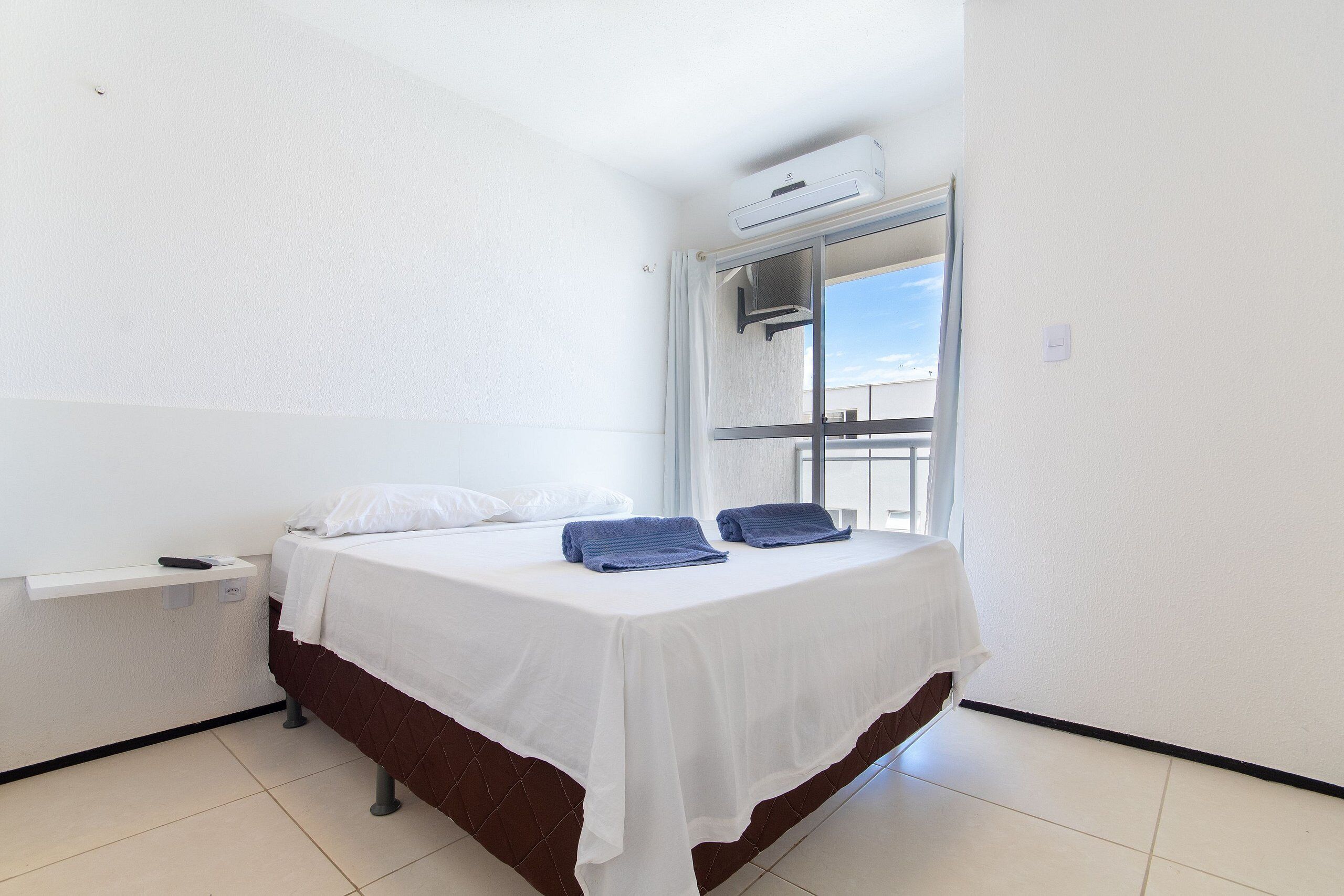 Una cama individual con ropa de cama blanca y marrón, una mesita de noche pequeña con un control remoto, y una ventana con vistas al cielo.