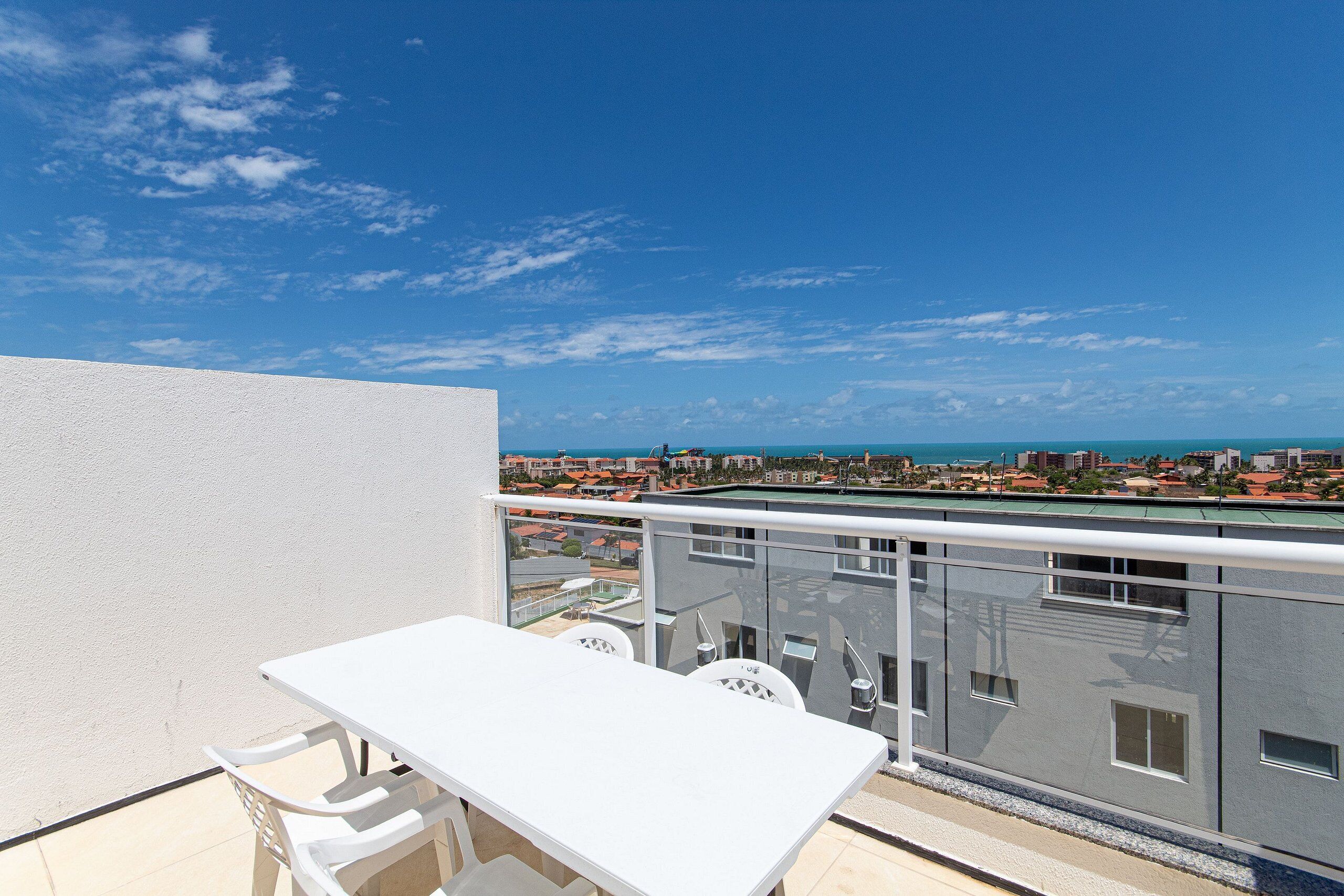 A balcony with a white table and chairs, overlooking a coastal cityscape with buildings and the sea.