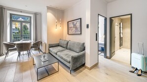 Una habitación de hotel moderna con sofá, mesa de comedor y sillas, un ventanal con vistas y un baño visible a través de una puerta abierta.