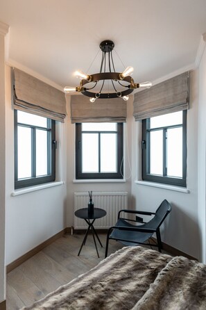 A corner room with a black chair, a small table, and a fur throw. There are three windows with blinds, a radiator, and a unique chandelier.