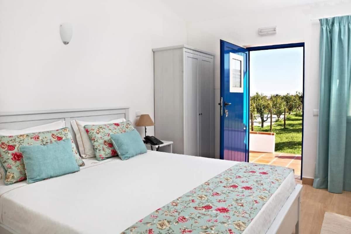 A bedroom with a bed, a nightstand, a wardrobe, and a view of a garden through an open door.