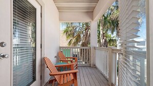 A balcony with wooden chairs and a table, a door with a window, and a view of palm trees. - Charming 3BR/2BA - 1/2 Block to Beach - Sleeps 8 from Rent On Padre (South Padre Island)