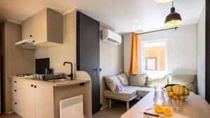 Une petite cuisine équipée avec une cuisinière, un évier et des placards, une petite table à manger avec des fruits, et un canapé dans une pièce lumineuse dotée d’un climatiseur et d’une fenêtre avec des rideaux.