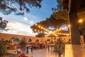 Uma área de pátio externo com mesas, cadeiras, luzes de festa e plantas em vasos.