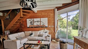 Un salon chaleureux avec un escalier en bois, un canapé d’angle, une table basse et une grande fenêtre donnant sur un paysage verdoyant.