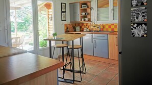 Une cuisine avec une table en bois, des tabourets de bar, un réfrigérateur décoré de photos et une fenêtre donnant sur un jardin.
