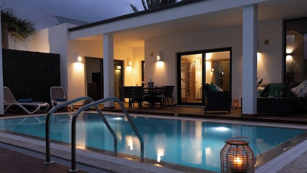 Una piscina con sillones de descanso y zona de comedor, rodeada por una casa moderna con ventanas amplias y un patio.