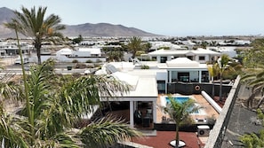 Una villa moderna con piscina, rodeada de palmeras y edificios blancos, con vistas a la costa.