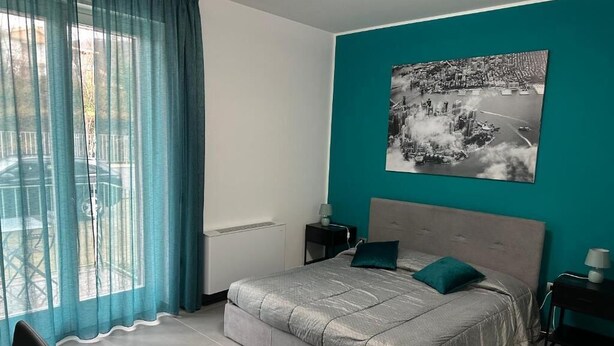 A bedroom with a bed, a nightstand, a window with curtains, and a cityscape painting on the wall. - Lotelito Apartment (Fisciano)
