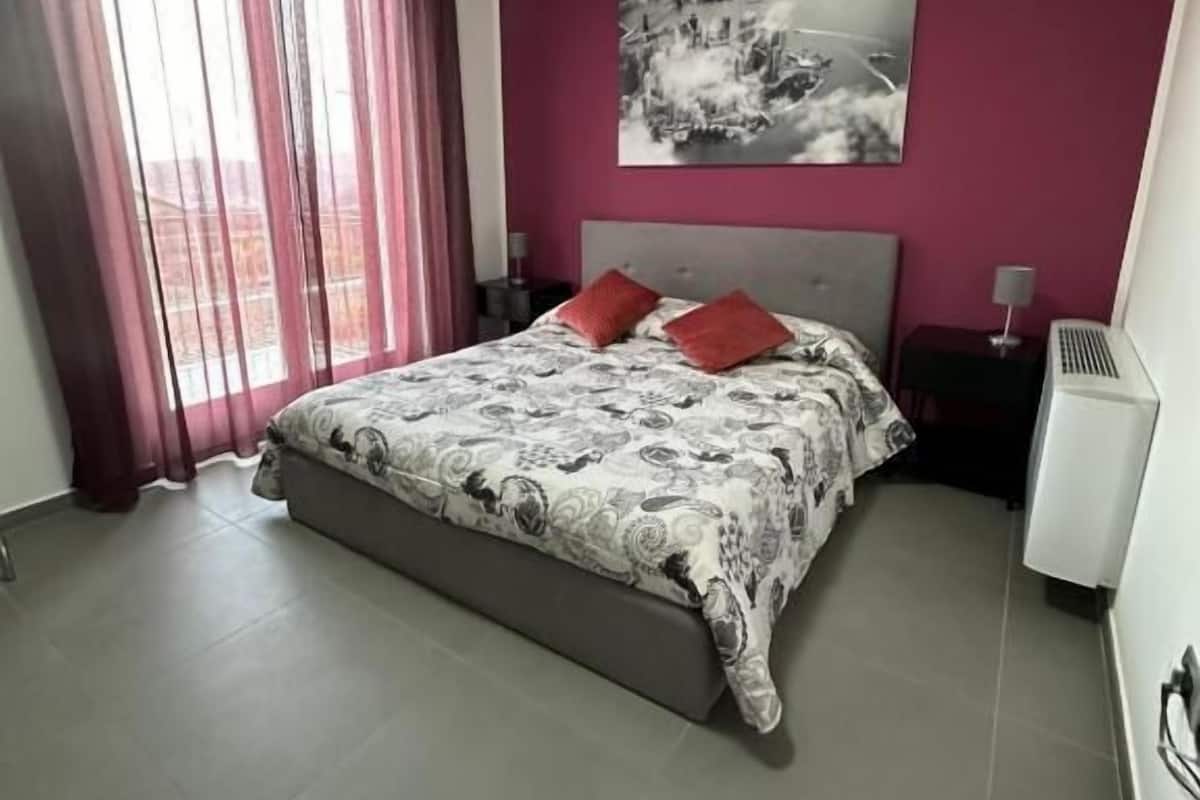 A bedroom with a bed, a nightstand, a heater, and a window with curtains.