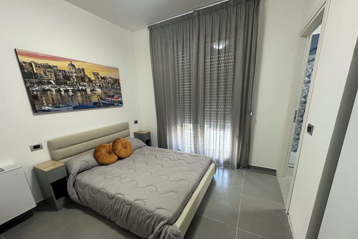 A bedroom with a bed, a nightstand, a wall-mounted painting, a window with curtains, and a door.