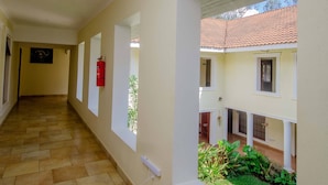 A corridor with tiled flooring, white walls, and windows overlooking a garden. - Amani Safari lodge  (Arusha)