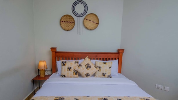 A neatly made bed with a wooden headboard, a bedside lamp, and decorative wall art. - Amani Safari lodge  (Arusha)