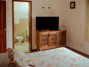 A bedroom with a bed, a wooden cabinet with a television, a bathroom visible through an open door, and a small framed picture on the wall. - House in a gated community with a pool – 300 meters from Taperapuã Beach (Porto Seguro)