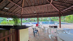 An outdoor pool area with a thatched roof, white plastic chairs, and a bar counter. - House in a gated community with a pool – 300 meters from Taperapuã Beach (Porto Seguro)