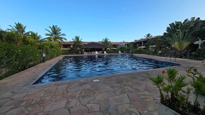 Una piscina rectangular con baldosas de piedra, rodeada de palmeras y un edificio de techo rojo.