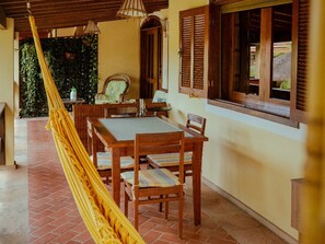 A room with a hammock, wooden furniture, a dining table, and a window with shutters. - House in a gated community with a pool – 300 meters from Taperapuã Beach (Porto Seguro)
