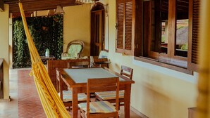 Un espacio exterior techado con zona de comedor, mesa, sillas, una hamaca y vistas a un jardín a través de una ventana.