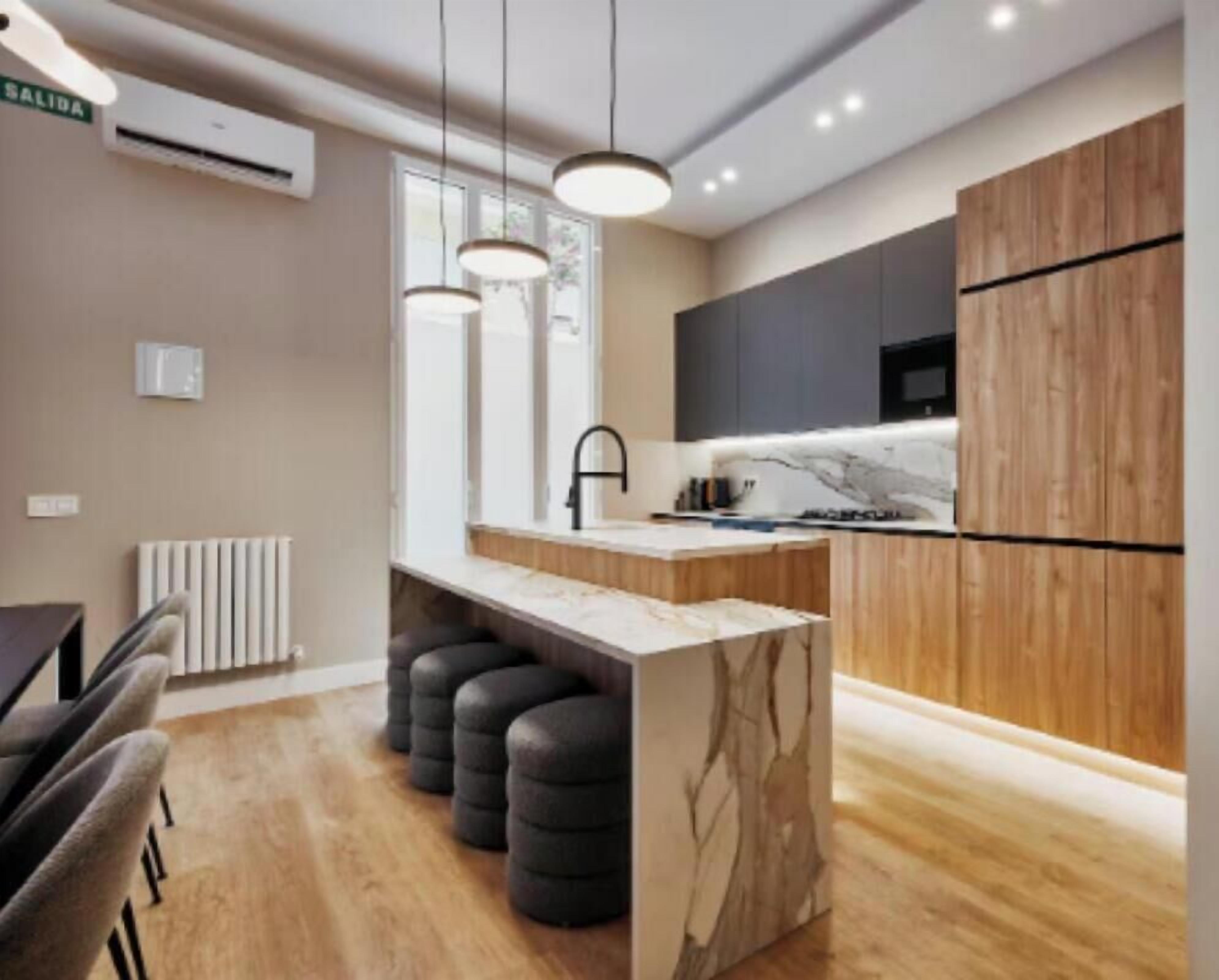A modern kitchen with a marble countertop, black bar stools, pendant lighting, and a built-in oven.