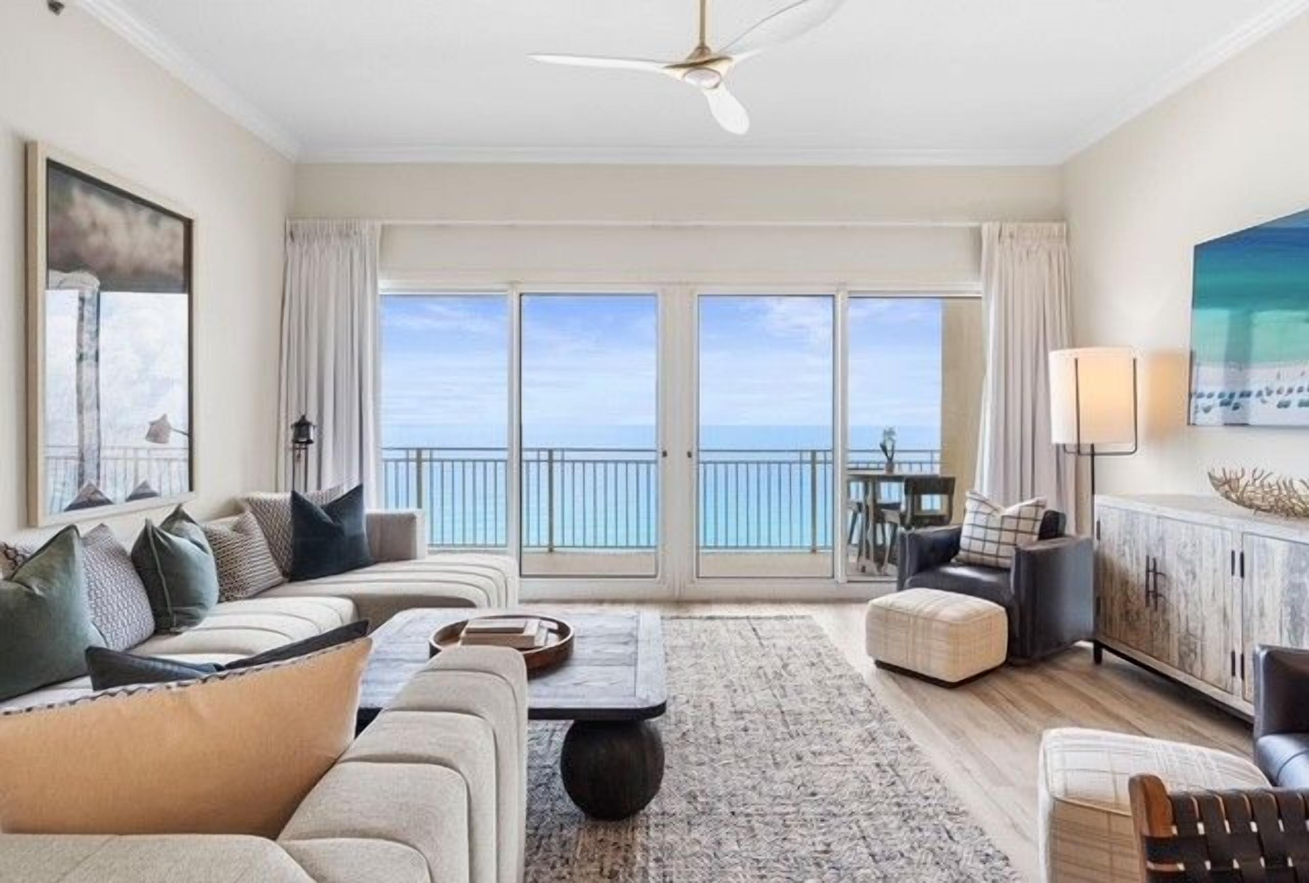 A spacious living room with a large window overlooking the sea, a ceiling fan, and a dining area with a view.