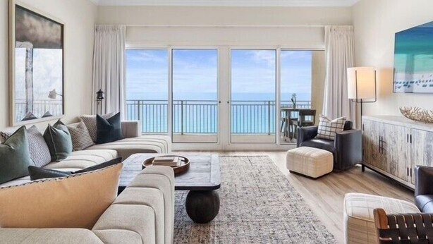 A spacious living room with a large window overlooking the sea, a ceiling fan, and a dining area with a view. - Sterling Beach 1202 Luxury Beachfront, Heated Pool, Walk in Shower, FREE Golf (Panama City Beach)