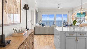 A spacious living area with a large window overlooking the sea, a modern kitchen with white cabinets, and a cozy seating area with cushions. - Sterling Beach 1202 Luxury Beachfront, Heated Pool, Walk in Shower, FREE Golf (Panama City Beach)