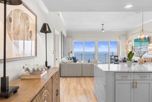 A spacious living area with a large window overlooking the sea, a modern kitchen with white cabinets, and a cozy seating area with cushions. - Sterling Beach 1202 Luxury Beachfront, Heated Pool, Walk in Shower, FREE Golf (Panama City Beach)