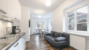 Een moderne keuken met roestvrijstalen apparatuur, een eethoek met een witte tafel en stoelen, en een grijze bank met uitzicht op een besneeerd landschap door grote ramen.