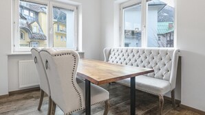 Een kamer met een houten eettafel, twee gestoffeerde stoelen, een knopenbank en grote ramen met uitzicht op gebouwen en bergen.