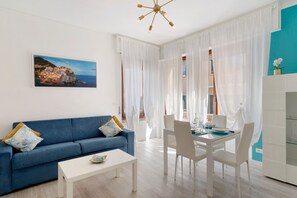 A modern living room with a blue sofa, white dining table, and chairs, a white coffee table, and a painting of a coastal scene. - Apartment 'A Casa Di Brenda' with Mountain View, Wi-Fi and Air Conditioning (Chiavari)