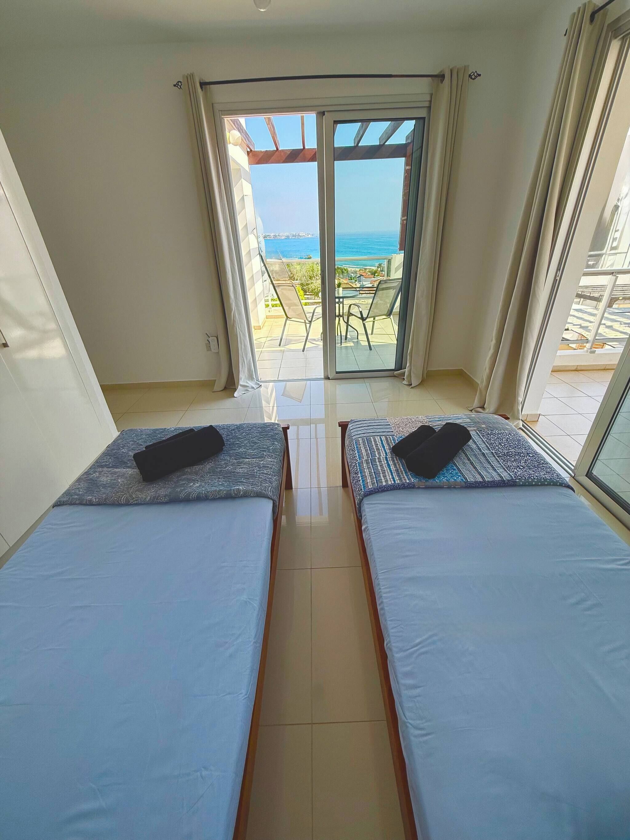 A room with two beds, a sliding glass door leading to an outdoor area with a view of the sea.