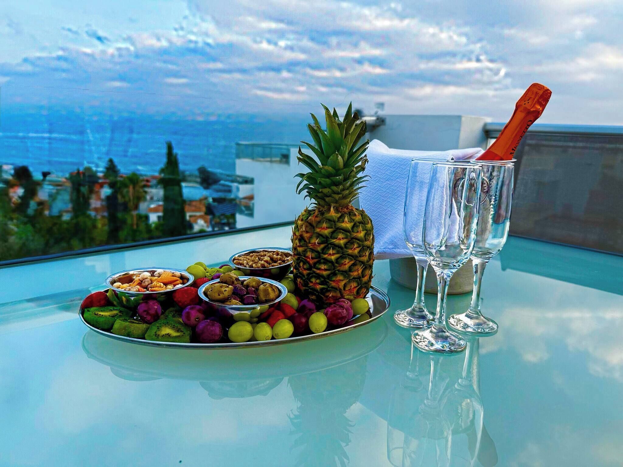 Een tafel met een fruitschaal, een ananas en twee champagneglazen op een balkon met uitzicht op de oceaan.