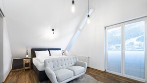 Ein Schlafzimmer mit Bett, Nachttisch, Sofa, großem Fenster mit Jalousien und Blick auf ein Gebäude.