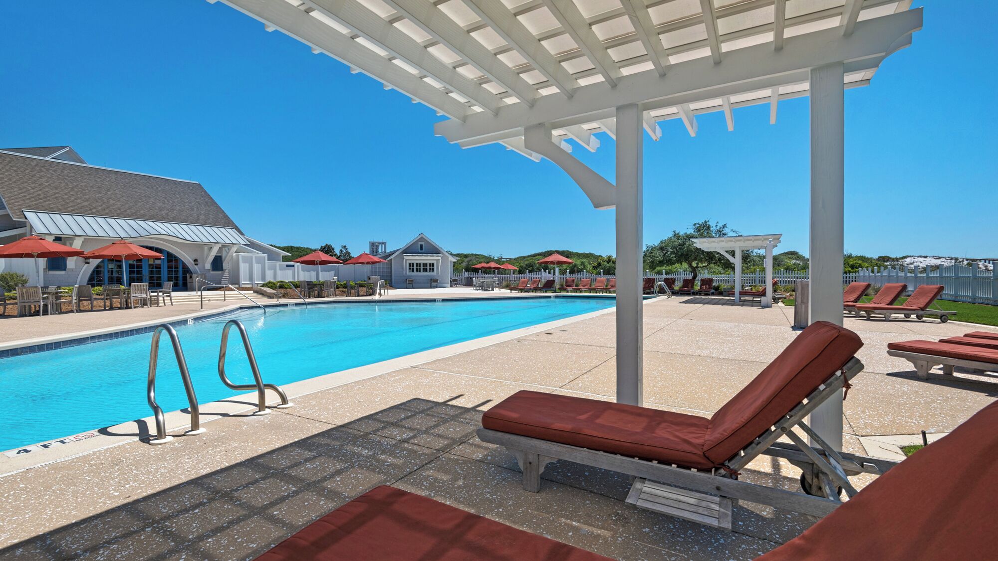 A swimming pool with lounge chairs and umbrellas, a covered seating area, and a building with a sloped roof in the background.