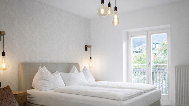 Un dormitorio con una cama grande, una mesita de noche de madera, un sillón tejido y vistas al exterior a través de un ventanal corredero.
