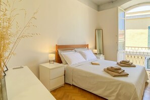 Ein Schlafzimmer mit Bett, Nachttischen, einem Spiegel, einem Fenster mit Blick und einer Vase mit Trockenblumen.
