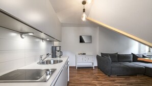 A modern kitchen with a built-in stove, a sink, and a coffee maker. There is a white cabinet with drawers and a white console table with a few items on it. A gray sofa is against the wall, and a wooden floor is visible. - Deluxe Apartment mit 57 m² für 2–4 Personen (Alpenresidenz (Bad Gastein))