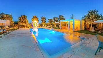 Una piscina esterna con un elemento d'acqua cristallina e azzurra, circondata da sedie a sdraio e ombrelloni, illuminata da luci soffuse.