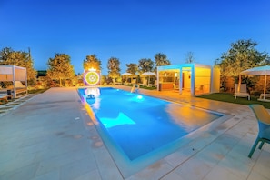 Piscina exterior con una característica de agua azul cristalina, rodeada de sillones de descanso y sombrillas, e iluminada con iluminación ambiental.