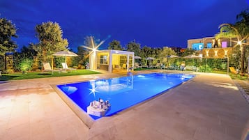 Una piscina con cerramiento de vidrio, rodeada de sillas de descanso y sombrillas, y una zona de patio bien iluminada.