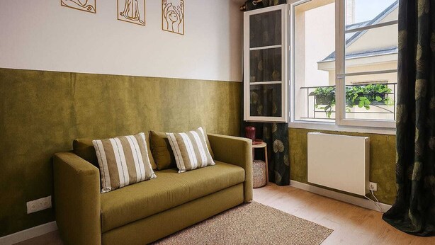 Un salon avec un canapé vert, des coussins à rayures, une petite table d’appoint, un radiateur et une fenêtre avec des rideaux.