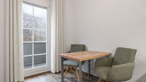 Una habitación con una mesa de madera, dos sillas verdes, una bombilla colgante, una ventana con cortinas y vistas a edificios.