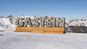 Un letrero con la palabra 'GASTEIN' sobre una plataforma de madera, con una montaña nevada al fondo.