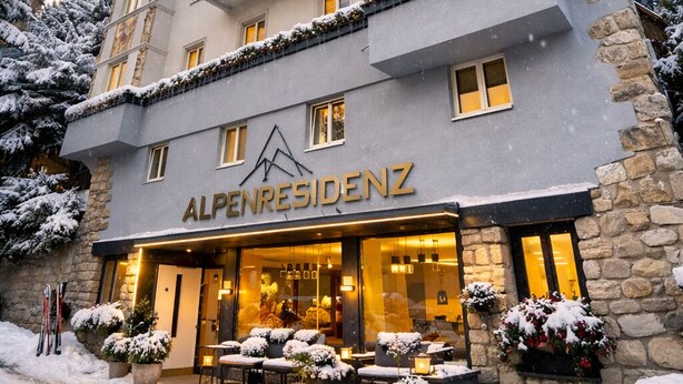Un hotel llamado Alpenresidenz con fachada de piedra, varios balcones y zona de estar exterior con plantas en macetas.