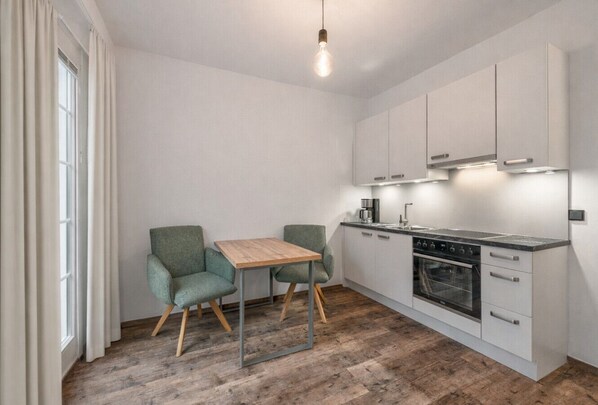 A compact kitchen with a wooden table and two chairs, a built-in oven, and white cabinets. - Modernes Studio-Apartment mit Balkon für 2 Personen (Alpenresidenz (Bad Gastein))