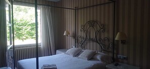 A four-poster bed with a metal headboard, two bedside lamps, and a view of trees through a window with curtains. - Cabanas, Casa Unica, on the beachfront (Cabañas)