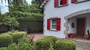 Un edificio blanco con contraventanas rojas, un arco de piedra y un pequeño jardín con setos y flores.