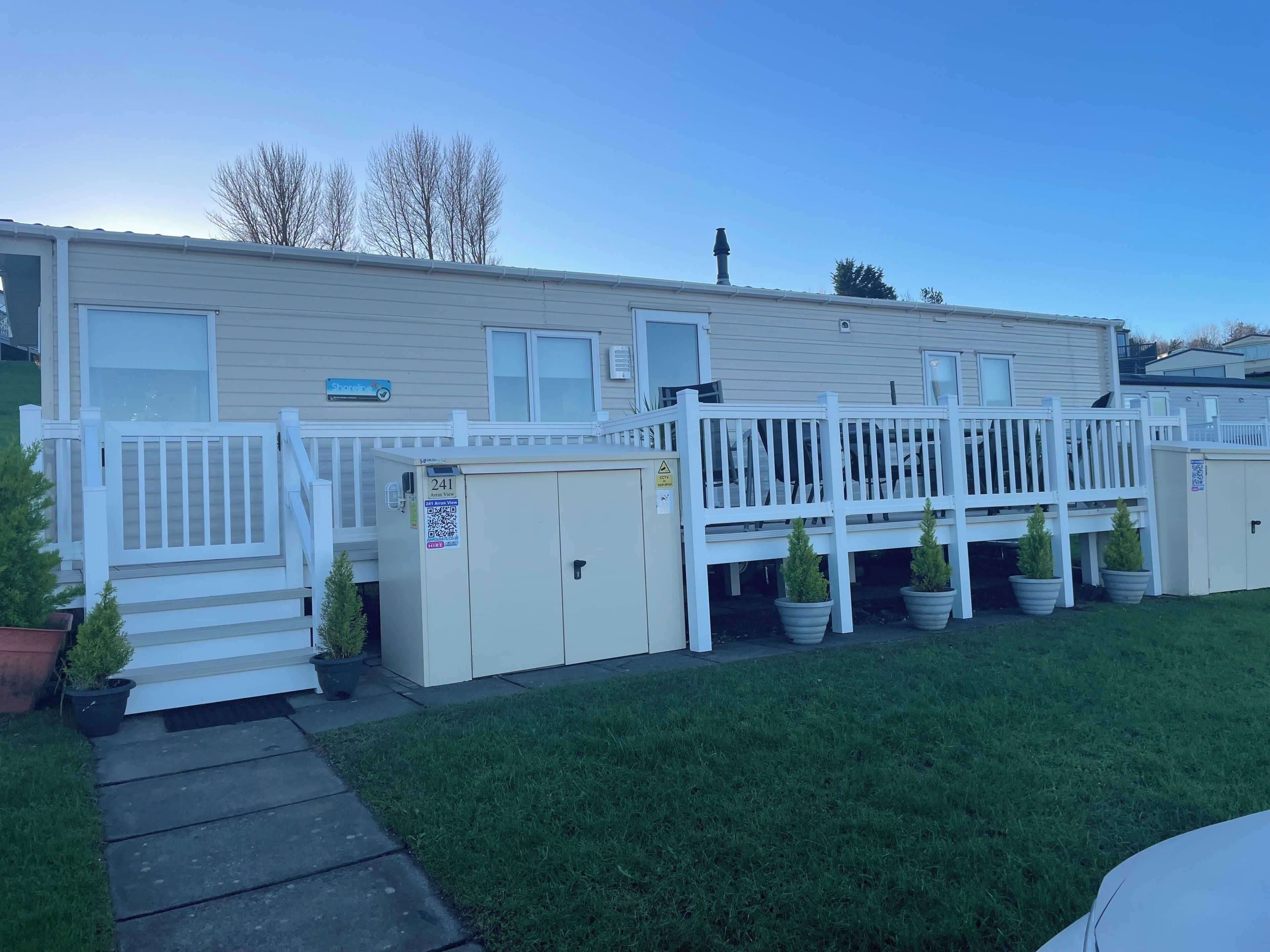 Une maison mobile dotée d’une terrasse clôturée, d’un garage et d’un petit jardin devant la maison agrémenté de plantes en pot.