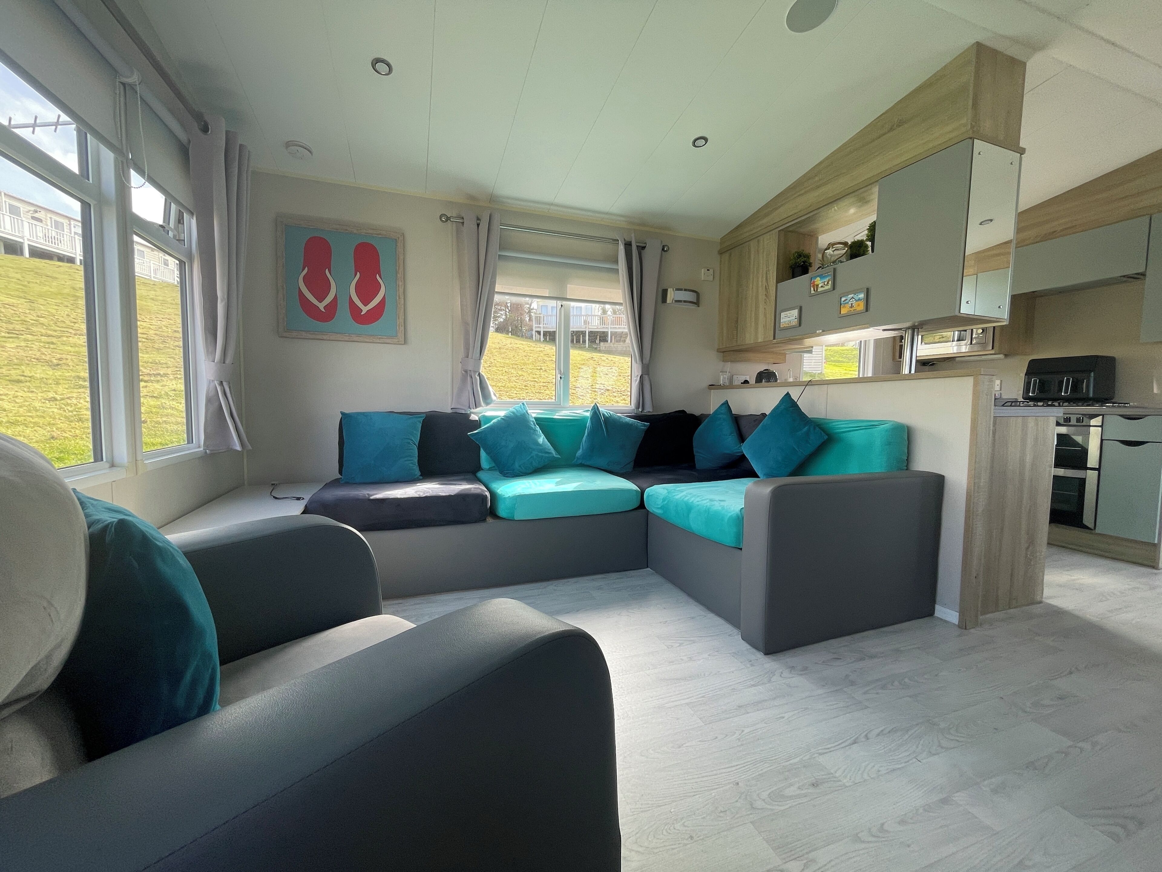 Ein modernes Wohnzimmer mit Ecksofa, einer Küchenzeile und großen Fenstern mit Blick ins Freie.